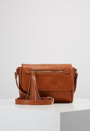 Sac bandoulière en cuir marron avec poche zippée à l'avant et détail pompon, posé sur une surface blanche sur fond gris clair.