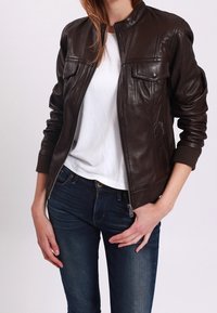 Brązowa skórzana kurtka typu bomber z podniesionym kołnierzem, z dwoma kieszeniami na piersi i zamkiem błyskawicznym z przodu. Noszona na białej koszulce i niebieskich dżinsach.