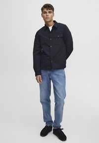 Navy blue, padded shirt jacket with button front, collar, single chest pocket, and black cuffs, paired with light blue jeans and black shoes.