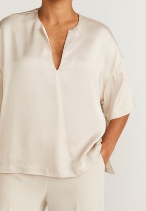 Persona que lleva una camiseta beige de manga corta y holgada con escote en V y pantalones beige a juego, de pie con una mano en el bolsillo.