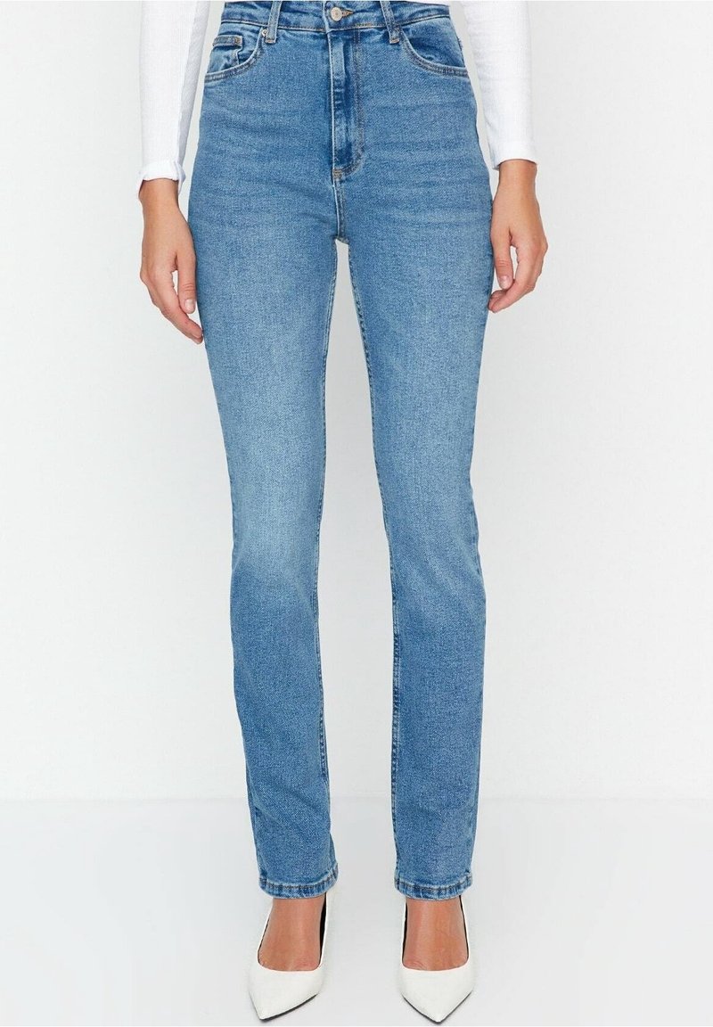 Femme portant un jean droit bleu clair taille haute avec un haut à manches longues blanc et des escarpins blancs à bout pointu.