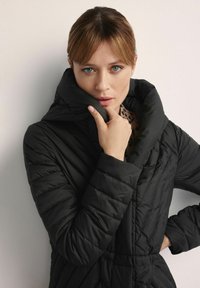 Czarna kurtka puchowa z wysokim kołnierzem, pikowanym wzorem i ściągniętym w Pasie. Modelka trzyma kołnierz jedną ręką.