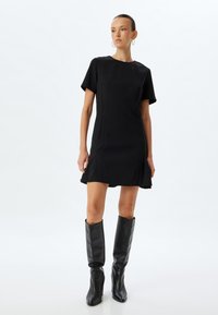 Schwarzes Kurzarmkleid mit tailliertem Oberteil und ausgestellt Rock. Glatter, matter Stoff; knielang. Kombiniert mit hohen schwarzen Lederstiefeln.