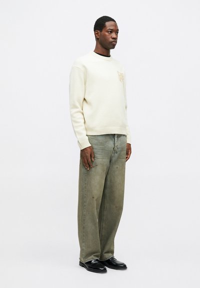 Férfi áll, világos krém színű pulóvert viselve, melyen egy kis tigris embléma található, széles, elhalványult zöld farmernadrágot és fekete elegáns cipőt visel, fehér háttér előtt.