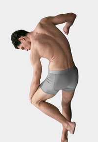 Uomo che indossa boxer grigi, piegato in avanti con una mano sul ginocchio e l'altro braccio piegato dietro, su uno sfondo neutro.