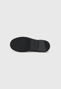 Semelle de chaussure en caoutchouc noire avec un motif de traction texturé, design plat, et une section de talon lisse, comportant une plaque de logo rectangulaire.