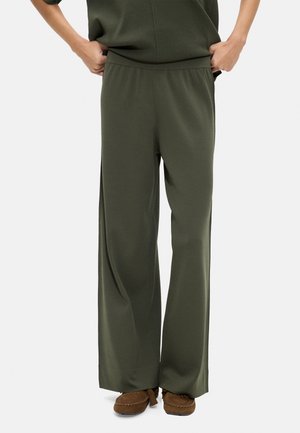 Osoba nosząca luźne oliwkowozielone spodnie z elastycznym paskiem w talii oraz brązowe buty w stylu mokasynów, ręce spoczywają blisko kieszeni.