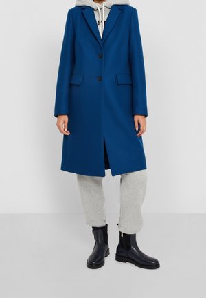 Personne portant un manteau bleu boutonné par-dessus un sweat à capuche gris, un pantalon de jogging gris et des bottines noires, debout devant un fond uni.