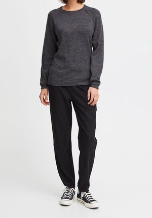 Femme portant un pull à manches longues gris foncé, un pantalon noir et des baskets noires, debout devant un fond blanc.