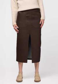 Bērnu ādas midi svārki ar priekšējo šķēlumu, divām lielām kabatām un redzamām saamšanas detaļām, kopā ar bēšiem slip-on apaviem.