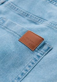 Světle modrá denimová látka s texturovaným povrchem a hnědou koženou štítkem, na kterém je vyznačeno jméno značky a "EST. 1985." Nedaleko zadní kapsy.