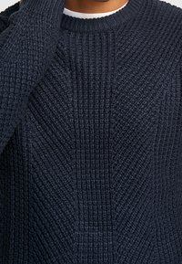 Marinblå stickad tröja med en strukturerad diagonalmönstring och rund halsringning, med ribbade detaljer längs kanterna. Mjuk, tjockt material.