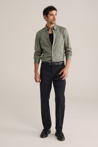 Camicia verde a maniche lunghe con bottoni sopra un top nero senza maniche, abbinata a pantaloni neri e scarpe nere. Tessuto liscio con una vestibilità classica.