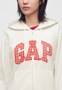 Personne portant un sweat à capuche blanc zippé avec un logo GAP à carreaux rouges et blancs sur la poitrine et un t-shirt rouge visible en dessous.