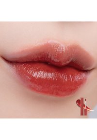 Labbra lucide in una ricca tonalità marrone rossastro con una texture liscia; un prodotto cosmetico mostrato nell'angolo con un applicatore a wand.