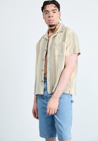 Camicia a maniche corte con bottoni a righe beige e grigie; taschino sul davanti, vestibilità rilassata. Indossata con pantaloni shorts in denim azzurro chiaro.