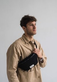 Black fabric fanny pack with a zippered closure, logo tag, and adjustable strap, worn across the chest by a person in a beige jacket.