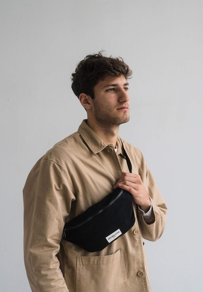 Black fabric fanny pack with a zippered closure, logo tag, and adjustable strap, worn across the chest by a person in a beige jacket.
