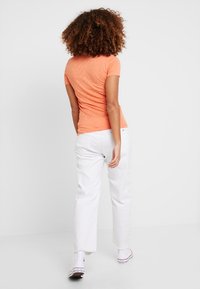 Vrouw met krullend haar, gekleed in een oranje shirt met korte mouwen, witte hoge taille broek en witte sneakers, staand tegen een plain witte achtergrond.
