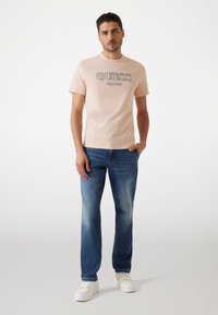 T-shirt de manga corta de color rosa claro con el logo "GUESS", combinado con jeans azules y zapatillas blancas, con un ajuste relajado y cuello redondo.
