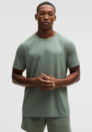 Mand står med hænderne foldet, iført en lysegrøn kortærmet skjorte og matchende shorts foran en ensfarvet baggrund.