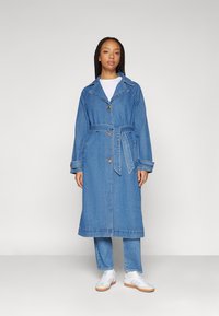 Denim trench coat gallér nélküli nyakkal, hosszú ujjakkal és övvel. Világoskék színű, látható varrásokkal és gombdíszítéssel.