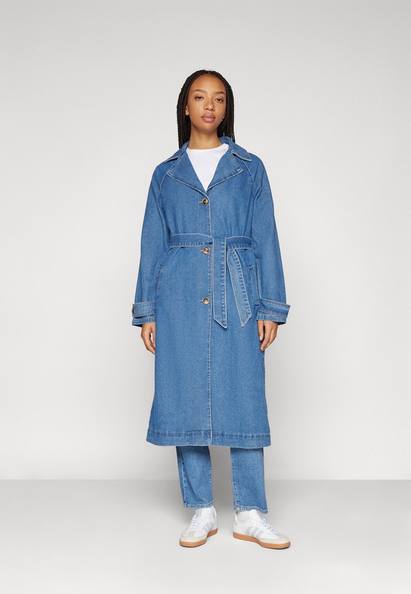 Denim trench coat gallér nélküli nyakkal, hosszú ujjakkal és övvel. Világoskék színű, látható varrásokkal és gombdíszítéssel.
