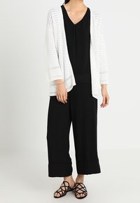 Vit, öppen stickad kofta över en svart topp och vida svarta byxor. Svarta sandaler med remmar på fötterna, vilket skapar en lager på lager, monokrom look.