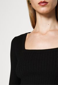 Femme portant un top noir à col carré côtelé, avec de longs cheveux raides et des boucles d'oreilles en or, lèvres avec du rouge à lèvres marron clair.
