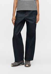 Άτομο που φοράει σκουρόχρωμα τζιν με φαρδιά παντελόνια, γκρι παπούτσια πεζοπορίας και μια μαύρη και λευκή ριγέ μπλούζα, στέκεται μπροστά σε απλό φόντο.