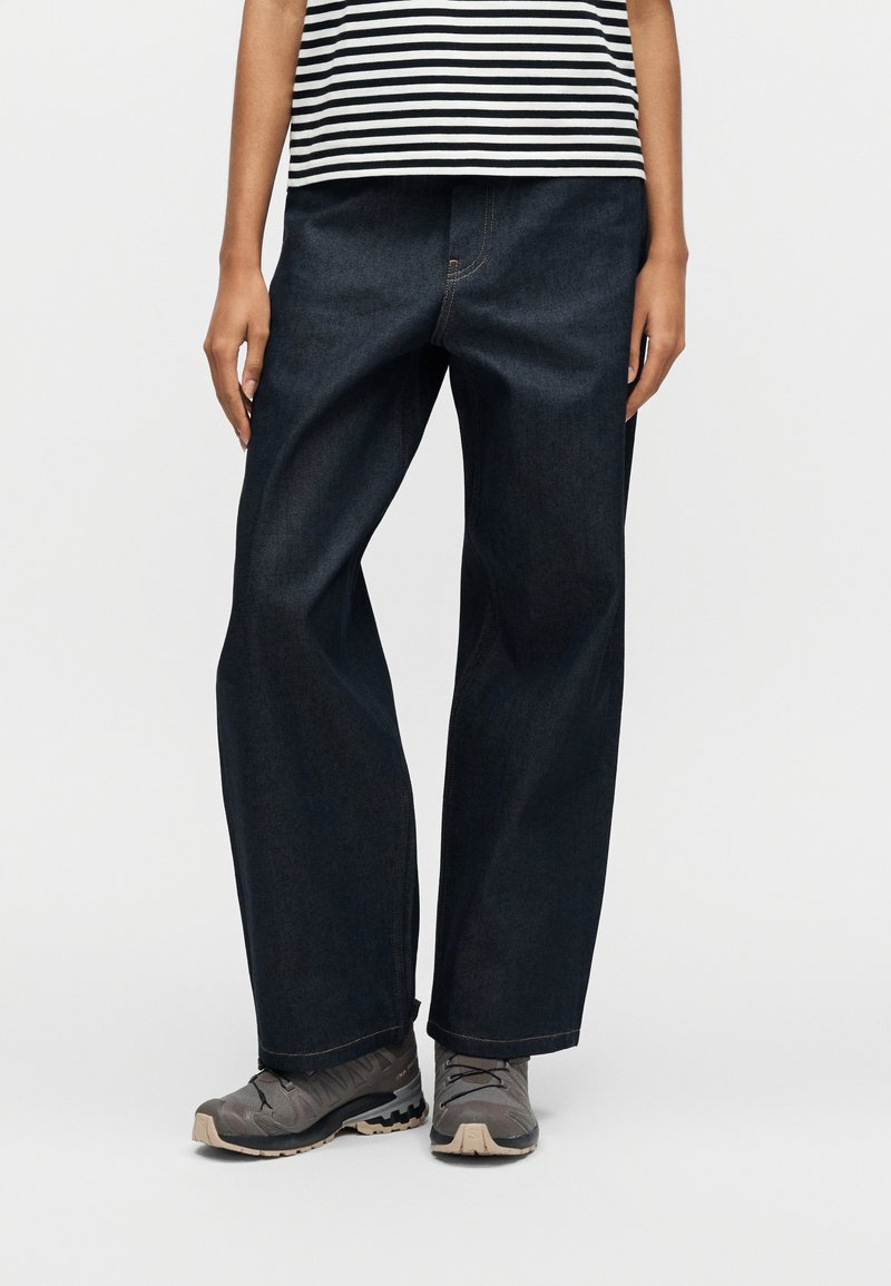 Person iført mørke, brede jeans, grå vandresko og en sort-hvidstribet skjorte, stående foran en ensfarvet baggrund.