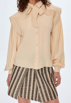 Blouse beige à manches longues avec col lavallière et détail plissé aux épaules, associée à une mini-jupe à rayures texturée taille haute.