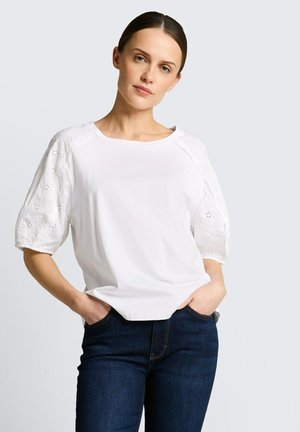 Vrouw met donker haar in een witte blouse met oogjespofmouwen en donkerblauwe spijkerbroek, staand met handen in de zakken tegen een effen achtergrond.