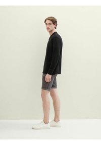 Cămașă neagră cu mânecă lungă și design cu guler, combinată cu șorturi gri și adidași albi, prezentând o ținută casual.
