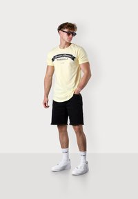 Helles gelbes T-Shirt mit schwarzem Grafikdruck, kombiniert mit schwarzen Denim-Shorts. Accessoires umfassen Sonnenbrillen und weiße Sneakers mit kurzen Socken.