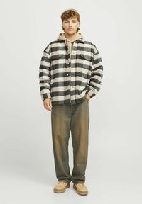 Camicia a maniche lunghe con motivo a quadri bianco e nero, indossata sopra un hoodie beige, abbinata a pantaloni verdi e scarpe beige. Look casual.