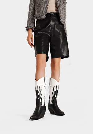 Pantaloni shorts in pelle nera abbinati a stivali da cowboy bianchi con dettagli di fiamme nere. L'outfit include una giacca grigia con texture.