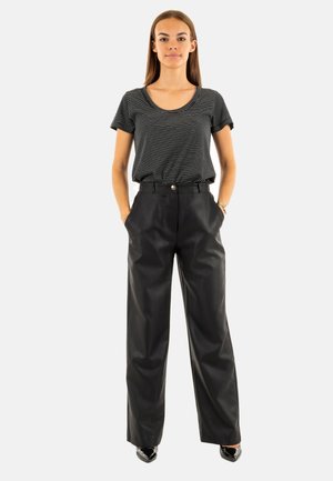 Jeune femme debout, les mains dans les poches, portant un tee-shirt rayé noir et blanc, un pantalon large taille haute noir et des talons noirs.