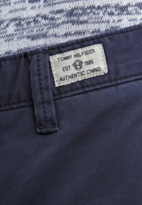 Námořnické modré chino kalhoty s hladkou texturou. Štítek uvádí "Tommy Hilfiger, Authentic Chino, Est. 1985." Obsahují čisté švy a jednolitý design.