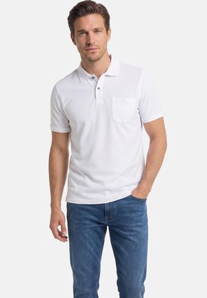 Homme portant un polo blanc à manches courtes avec poche poitrine et un jean bleu, debout une main dans la poche, sur un fond uni.
