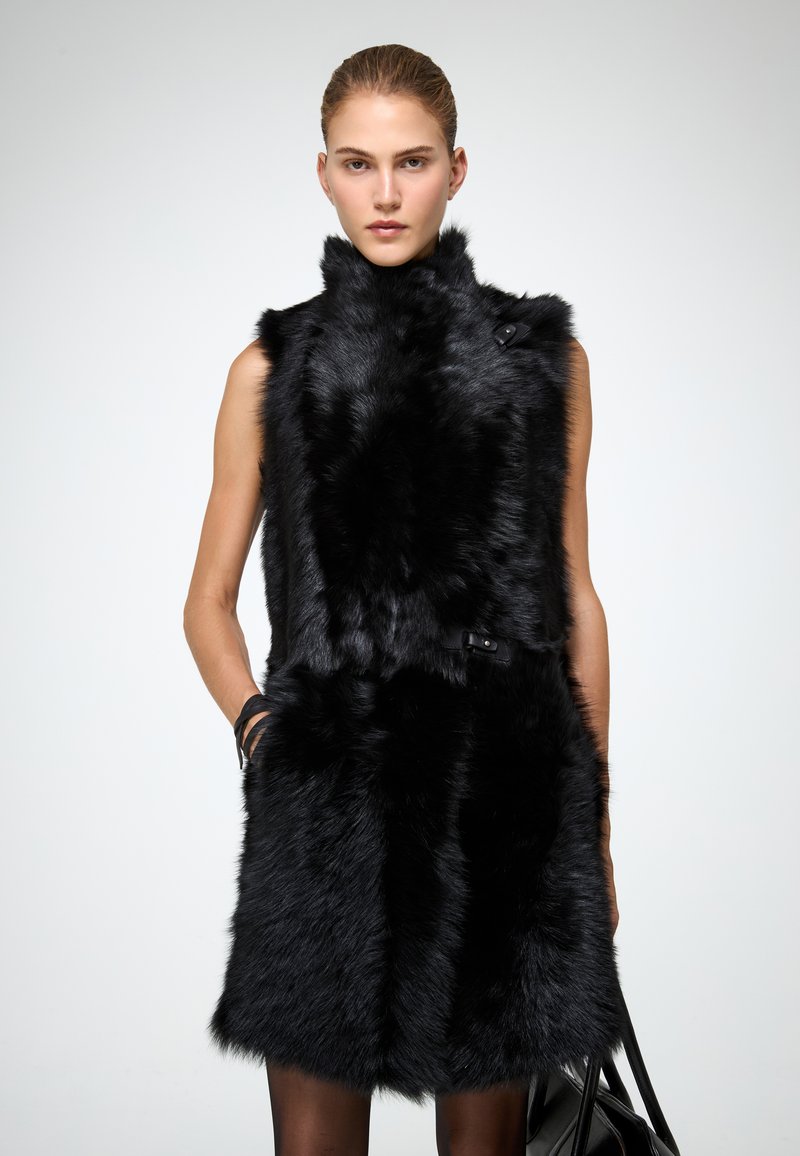 Juodas dirbtinio kailio liemenė su aukštu apykakle, dvigubais mygtukais ir glotnia siluetu. Lygi tekstūra, nukirptas apatinis kraštas ir be rankovių.