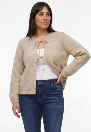 Kvinde iført beige strikket cardigan bundet på to steder over en hvid top og blå jeans, stående med den ene hånd på hoften.
