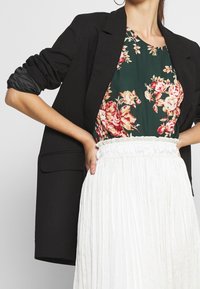 Černé sako, květinový zelený top s červenými a žlutými vzory, bílá skládací sukně s elastickým pasem. Hladké tkaniny a propracovaný design.
