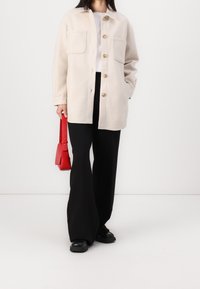 Light beige button-up jacket with two front pockets, paired with black wide-leg pants and a small red handbag. Smooth fabric texture.