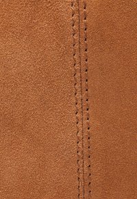 Brązowy zamsz o miękkiej teksturze, z wyraźnym szwem wzdłuż jednej strony, z blisko rozmieszczonymi detalami przeszycia.