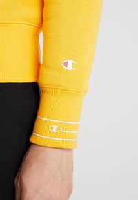 Sweat-shirt jaune avec poignets côtelés ; logo blanc sur la manche et bandes d'accent. Texture de tissu lisse.