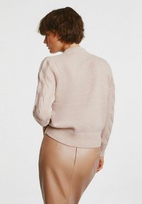 Jasnoróżowy sweter z dzianiny z prążkowanym wykończeniem i rękawami w splot warkoczowy, noszony na beżowych skórzanych spodniach, prezentujący teksturowany wzór.