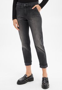 Femme portant un jean noir droit avec revers, doté de poches plaquées à l'avant et de mocassins noirs, se tenant avec une main dans la poche.