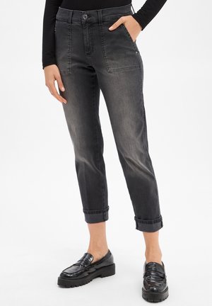 Femme portant un jean noir droit avec revers, doté de poches plaquées à l'avant et de mocassins noirs, se tenant avec une main dans la poche.