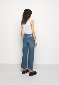 Jean large bleu en denim, taille haute avec un design à cinq poches. Associé à un haut sans manches blanc et des chaussures noires.